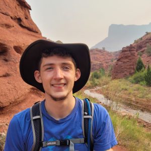 Casual outdoor portrait of Nick Christensen, wearing a hat and backpack, standing on a trail near a red rock formation and a small stream.