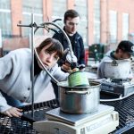 Student looking at cookstove prototype while other students observe or work on laptop