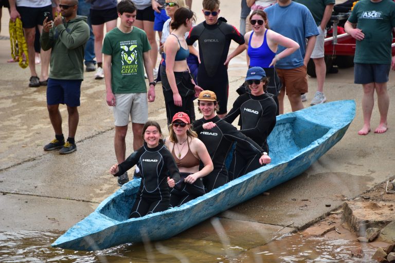 Four students sit in concrete canoe with other standing behind.