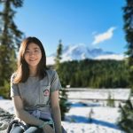 Woman sitting in snowy park with trees and mountains in the background.