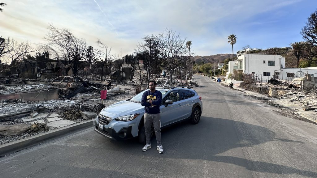 Man stands by car surrounded by wildfire wreckage.