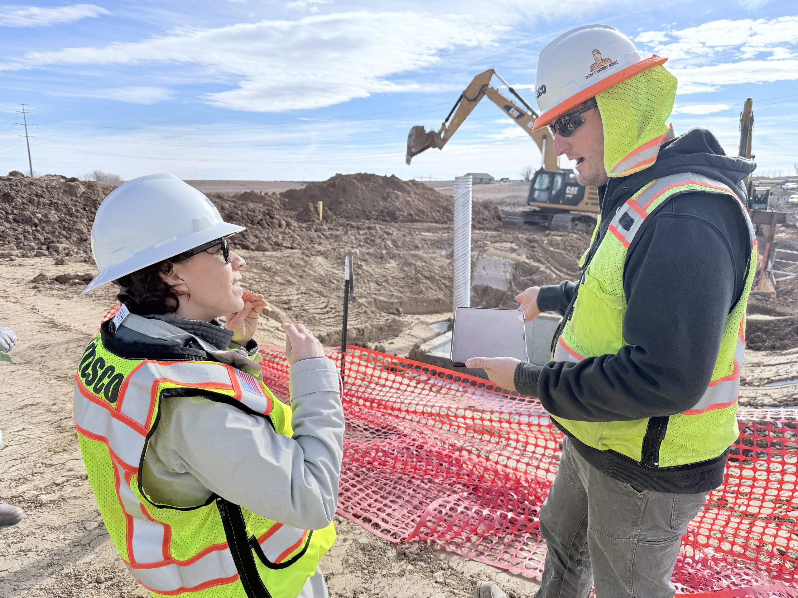 Two individuals in safety gear talk with heavy machinery in background.