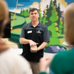 Man in black shirt with green mural background speaks to students.