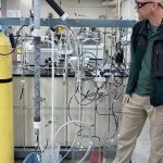 Man in safety googles inspects tall beakers of fluid.