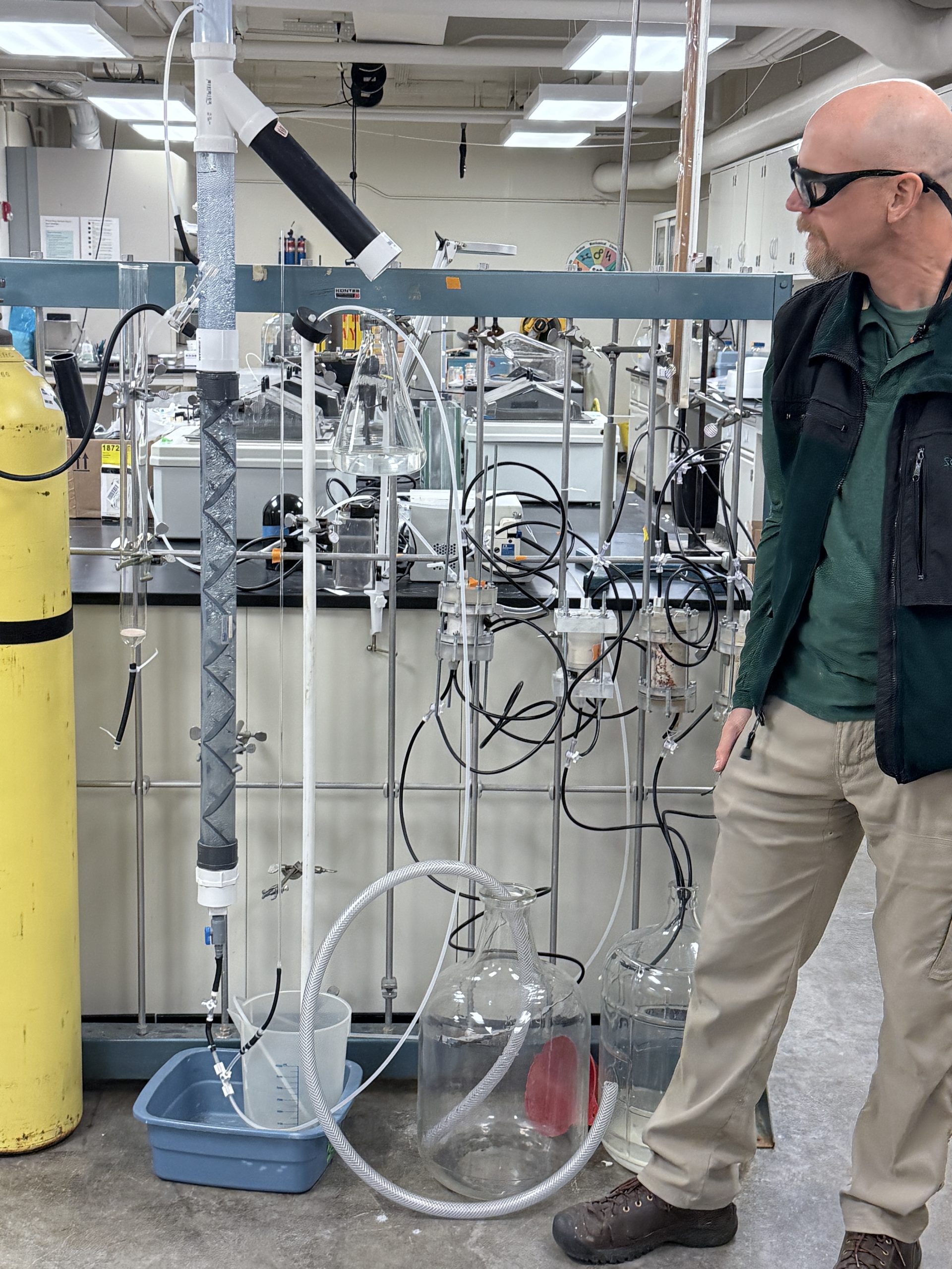 Man in safety googles inspects tall beakers of fluid.