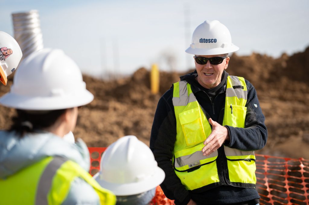 Man in hardhat and vest speaks to group at construction site.