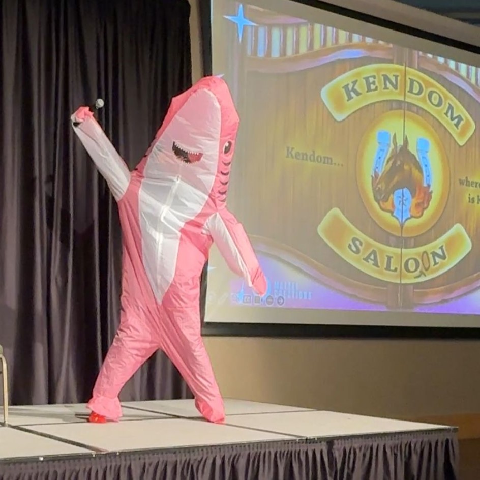 Person in pink inflatable shark costume holds microphone in the air.