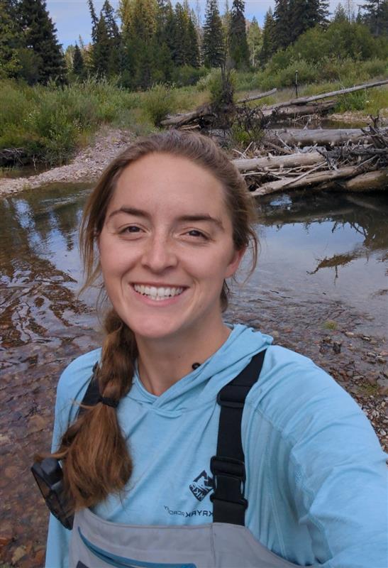 Woman in waders stands with creek in background.