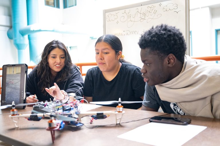 Three students sitting at table looking at their drone class project