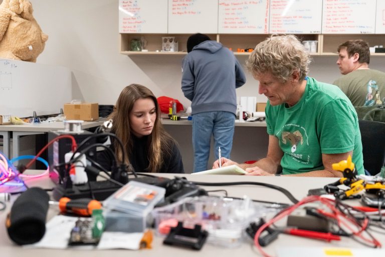 Female student and volunteer engineer working together in the lab, with equipment on table in front of them, with students in background