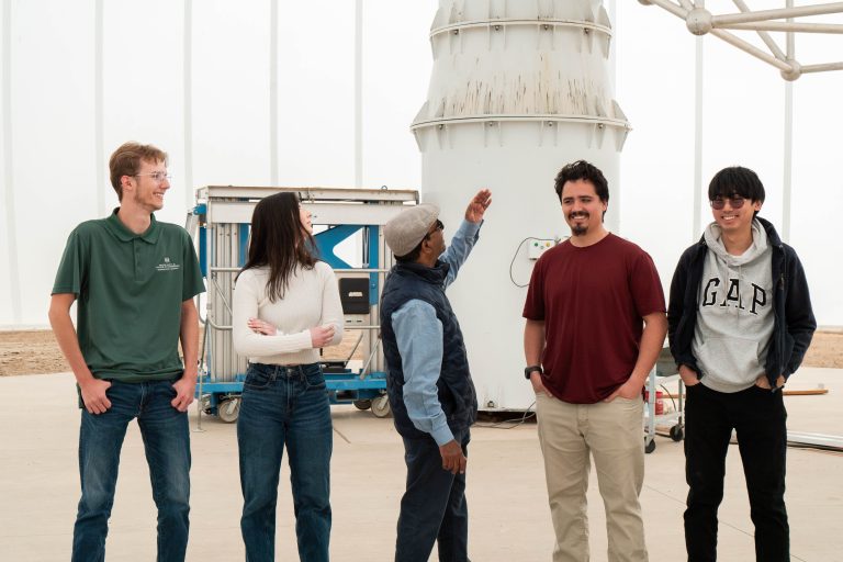 Four students stand with professor who is pointing to very large satellite