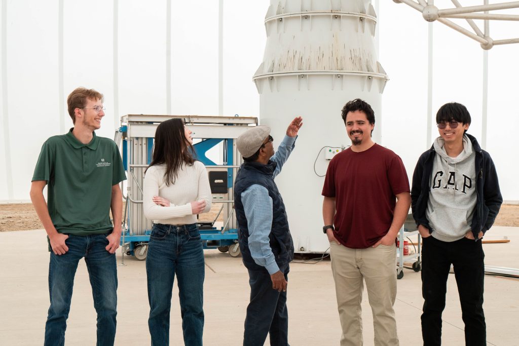 Four students stand with professor who is pointing to very large satellite