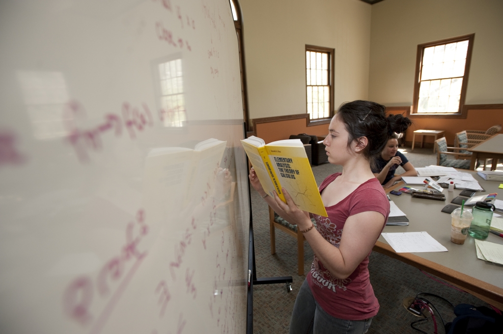 Student writes equation on white board while holding a book