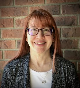 A professional photograph of Dr. Jennifer Mueller against a brick wall background.