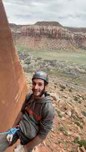 A photograph of Kyler Howard on a climbing excursion. He is attached to a canyon wall and wears climbing gear. Red canyons and desert are visible behind him.