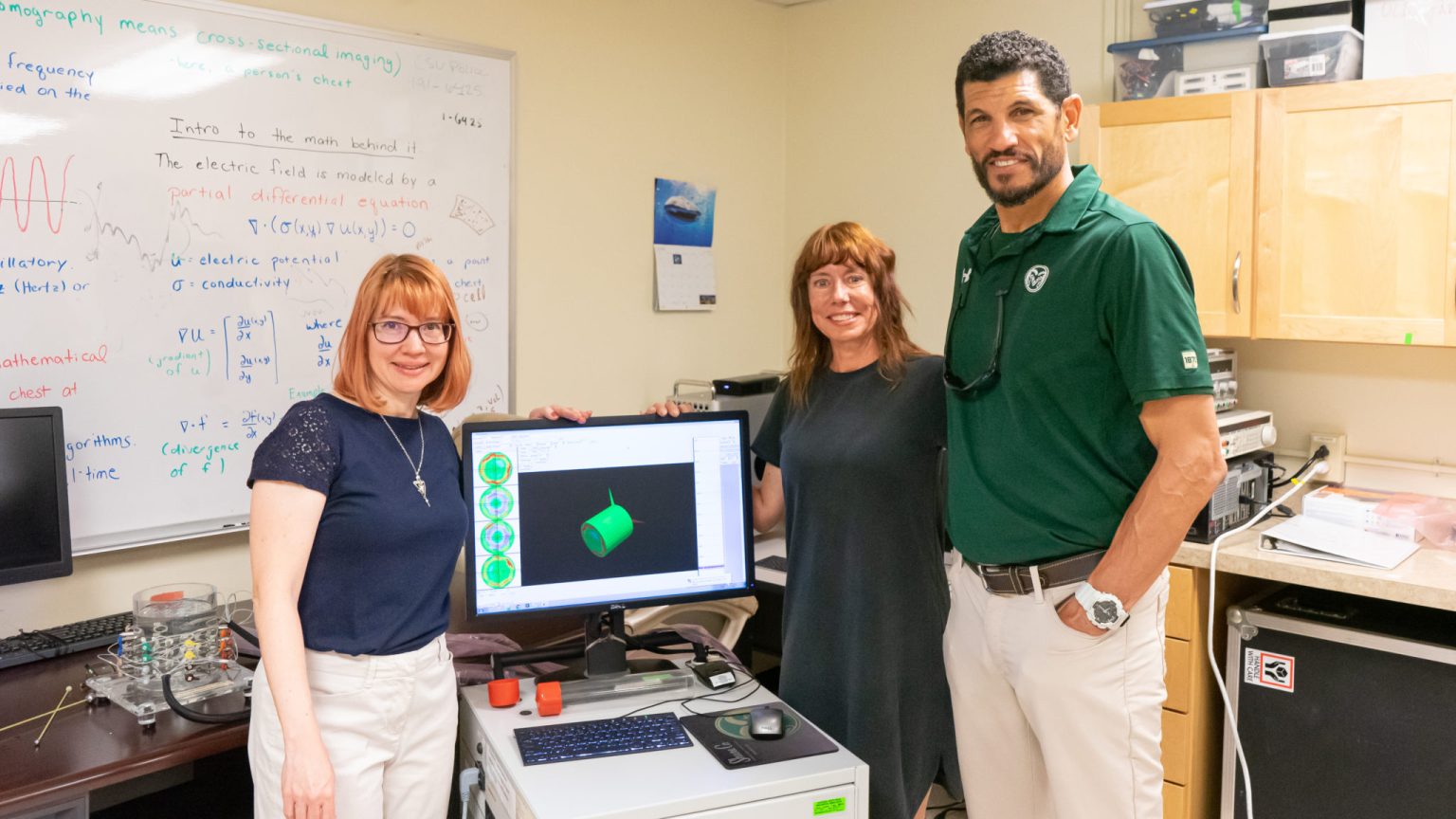 Dr. Jennifer Mueller, Kim Norvell, and Jay Norvell pose with an EIT system. A reconstruction of a human torso is displayed on the screen.