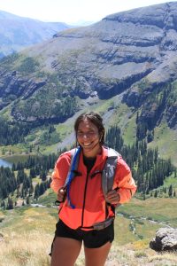 A photograph of Natalie Wijesinghe on a hike. SHe wears an orange blazer and poses in front of a mountain vista with trees and a lake.
