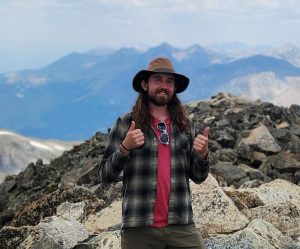 Trevor stands in front of a mountain view. He is wearing a sun hat and giving a double thumbs up.