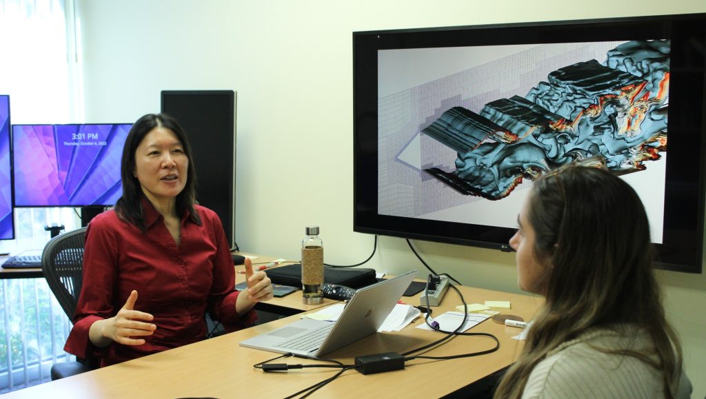 Dr. Xinfeng Gao sits at her desk with a female student. A colorful graphic projected on a TV screen plays behind them.