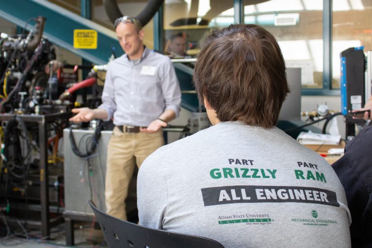 A student sits in front of a teacher lecturing. The back of the student's shirt reads, "Part Grizzly. Part Ram. All Engineer. Adams State University, Colorado State University Department of Mechanical Engineering.