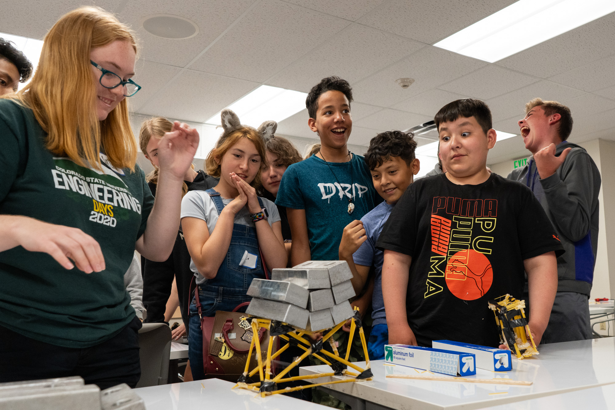 A group of student surround a bridge made out of spaghetti with weights on top of it.