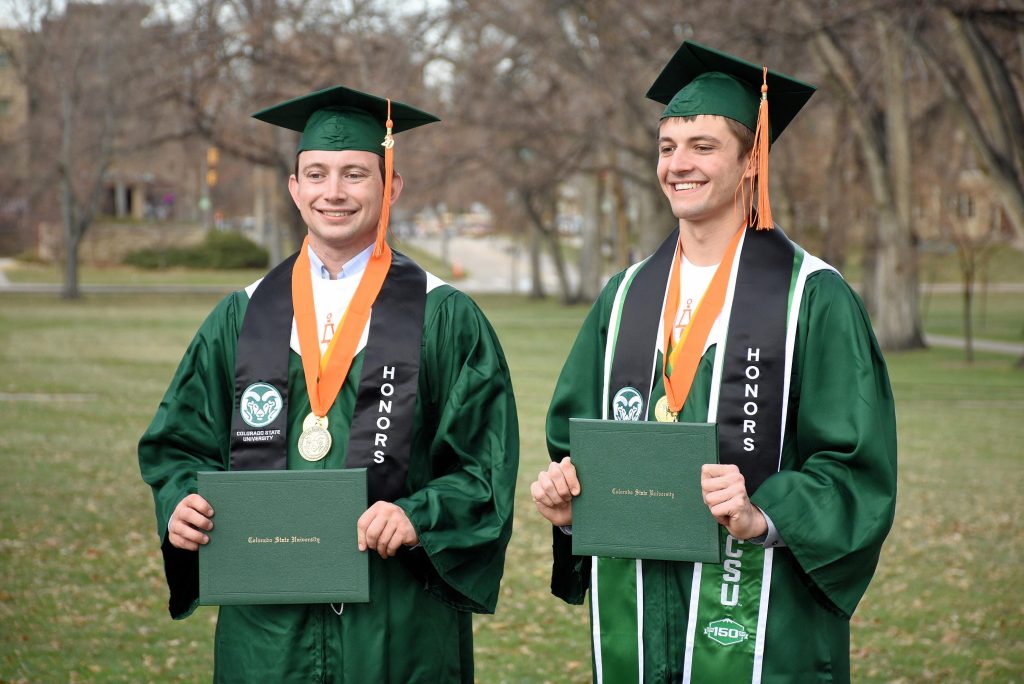 Two male students stand smiling in their Colorado State University regalia with their diplomas.