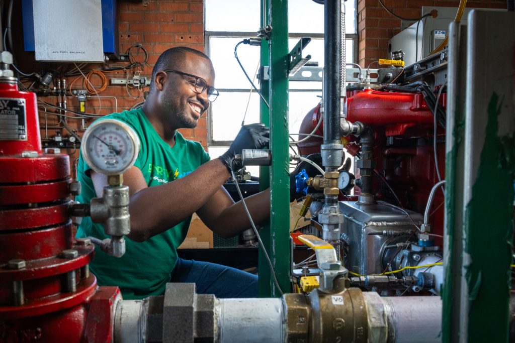 A male student works on colorful machinery. He's surrounded by colorful pipes, gauges, and wires.