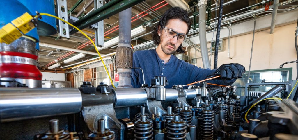 A male student uses a hand crank to adjust components on an engine.
