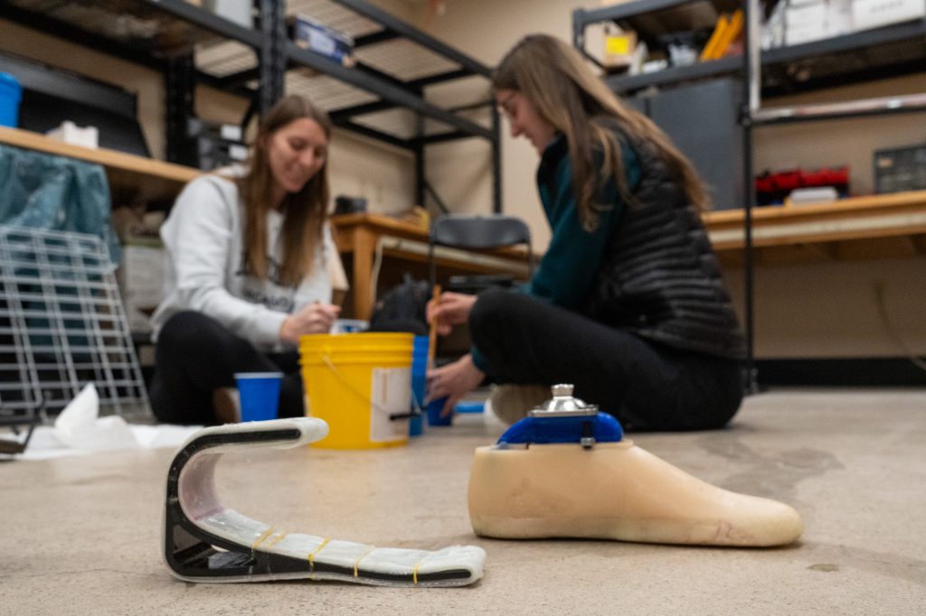 Two female students sit in the background of the photo stirring liquid in colorful buckets. In the foreground are two prosthetic feet prototypes. The first is very simplistic, and the one on the right resembles more of a human foot.