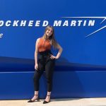 Kori Eliaz standing and smiling in front of a blue wall with the Lockheed Martin logo on it.