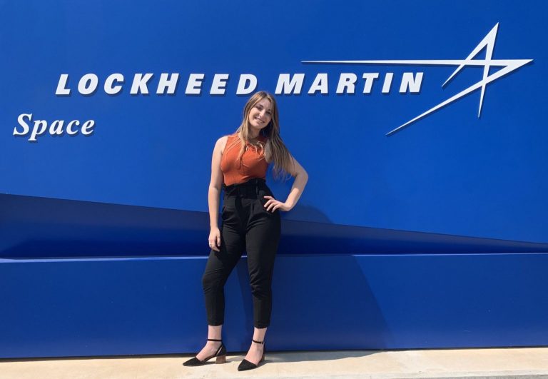 Kori Eliaz standing and smiling in front of a blue wall with the Lockheed Martin logo on it.