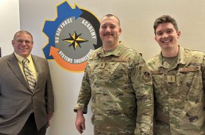 Jeremy Daily is a white man with short hair wearing glasses and a formal suit. Behind him is a crest on the wall that reads “US Air Force Academy Systems Engineering.” Maj. Trae Span is a white man with a short haircut and a mustache wearing his uniform. To the right of Span is 2nd Lt. Gabe Salinger, a young white man with brown hair also wearing his uniform. They are smiling at the camera.