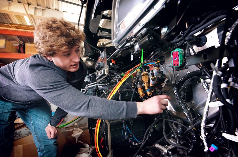 Jake Jepson is pictured bending over with one hand on his knee while the other gestures at the internal components of a heavy truck’s cab that were removed to be used as a testbed. A headlight from the cab testbed is in the at the top of the image with various wires, pipes, and plugs underneath. Jepson has on a gray sweater and blue jeans. He has curly dark blond hair and is looking toward the wires.