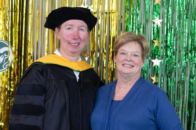 Sandra “Sandy” Dawson has on her doctoral regalia and is standing next to Ann Batchelor. Both are white women smiling. The background is strands of shiny party strings and stars in CSU colors."