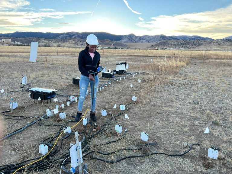 A women researcher in a hardhat with a wired network at her feet and mountains in the background.