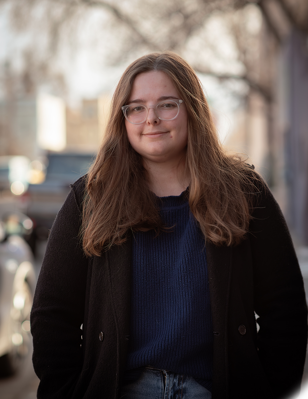 A woman in a light coat stands on a sidewalk with cars in the background.