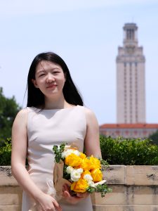A woman in a dress stands in front of a building holding flowers.  