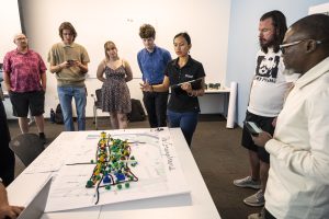 Seven people stand around a map with legos and other crafts material built to symbolize a cityscape. A young woman in the middle is speaking and gesturing.