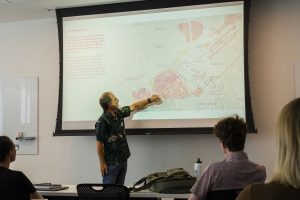 A man gestures at a projected map in front of a classroom.