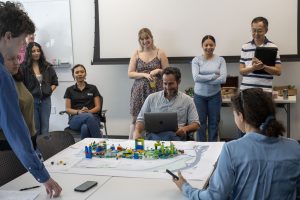 A group of people gather around a map with LEGO and other crafts on top depicting a mockup of a city. They are laughing. 