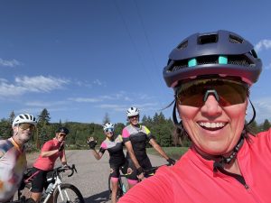 A woman in a bike helmet takes a selfie with four other cyclists smiling and waving in the background. 