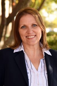 A woman smiles while wearing a suit jacket and standing outside near a tree.