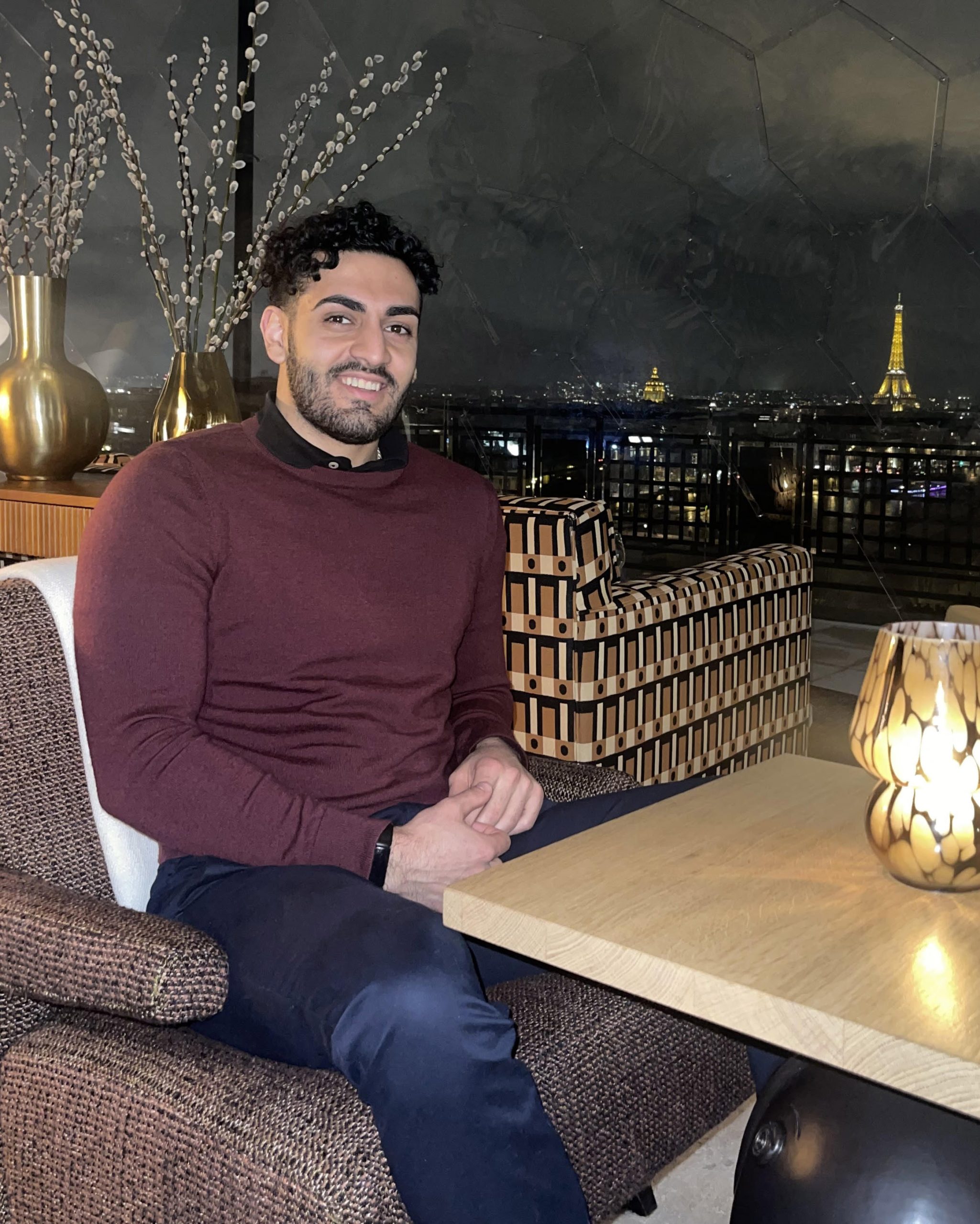 A man in nice casual clothing sits at a restaurant with the eiffel tower in the background.