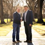 Two men in business casual clothing shake hands in the CSU Oval, on a sidewalk lined with trees.