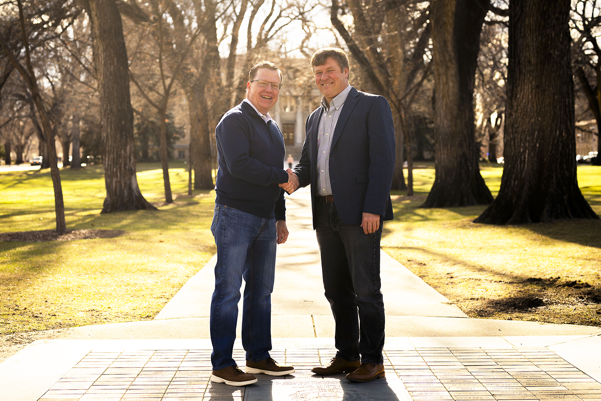 Two men in business casual clothing shake hands in the CSU Oval, on a sidewalk lined with trees.