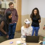 Six people gather around a laptop with the one in the middle sitting and using VR Goggles with hand controls.