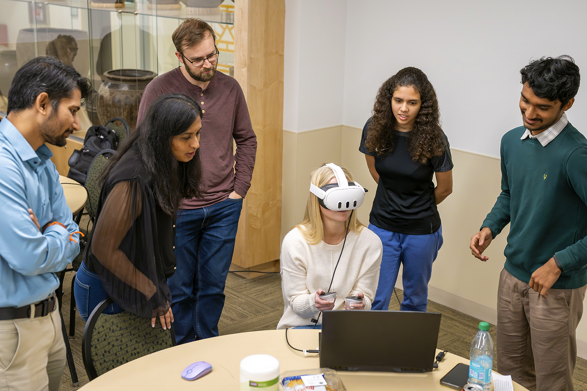 Six people gather around a laptop with the one in the middle sitting and using VR Goggles with hand controls.