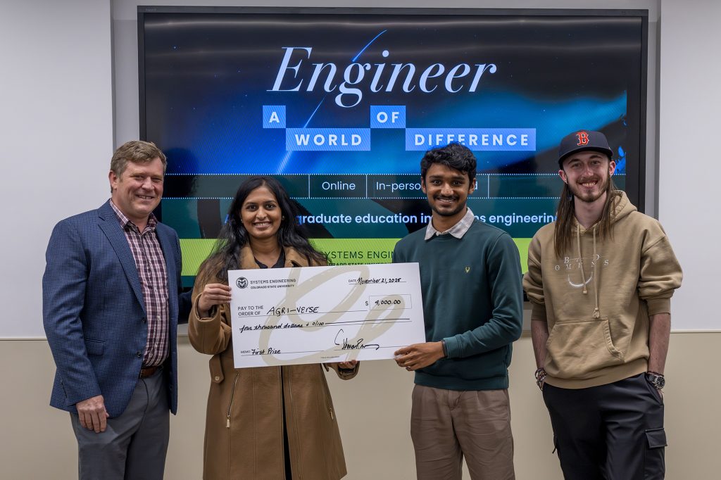 Four people stand in front of a tv screen with the words “engineer a world of difference” on it. The two in the center hold a giant fake check with $4000 listed on it.