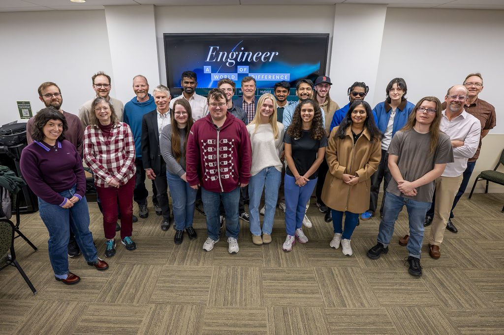 Twenty-two people stand in front of a TV with the text: “Engineer a world of difference” on it.
