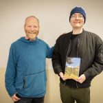A man in a sweatshirt stands next to another younger man in a beanie hate holding a book.